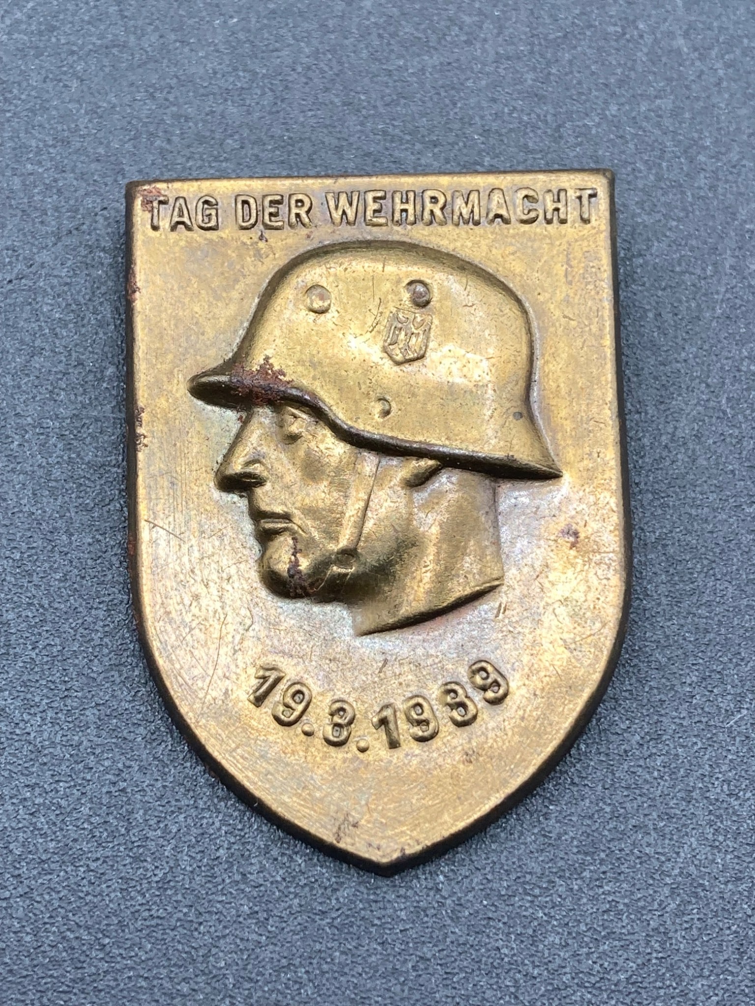 1939 German Day Of The Wehrmacht Tinnie Badge: This item is a 1939 German Day Of The Wehrmacht tinnie, featuring a raised profile of a soldier wearing a helmet with an insignia. The piece includes the date, 19.3.1939, inscribed below the helmet an