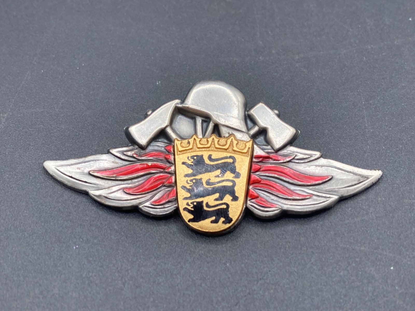 WWII German Baden-Württemberg Firefighting Cap Badge: This WWII-era German cap badge represents the firefighting services of Baden-Württemberg. It features a detailed design with two crossed axes, a firefighter helmet, and the three black lions of the B