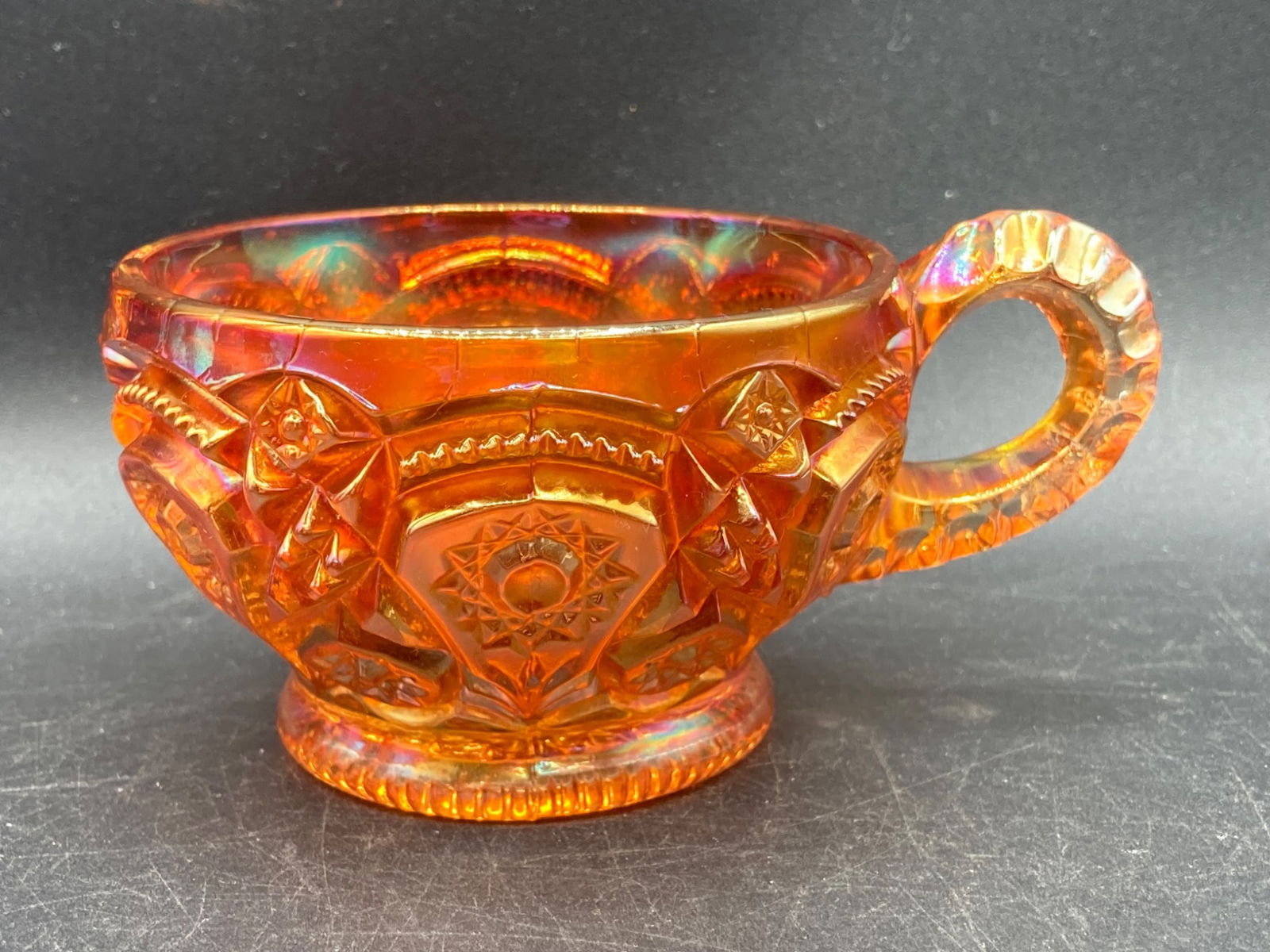 Carnival Glass Marigold Heirloom Tea Cup: This is a marigold-colored carnival glass tea cup featuring an heirloom pattern with intricate detailing. The cup measures approximately 3.5 inches in diameter and has a distinct iridescent finish cha