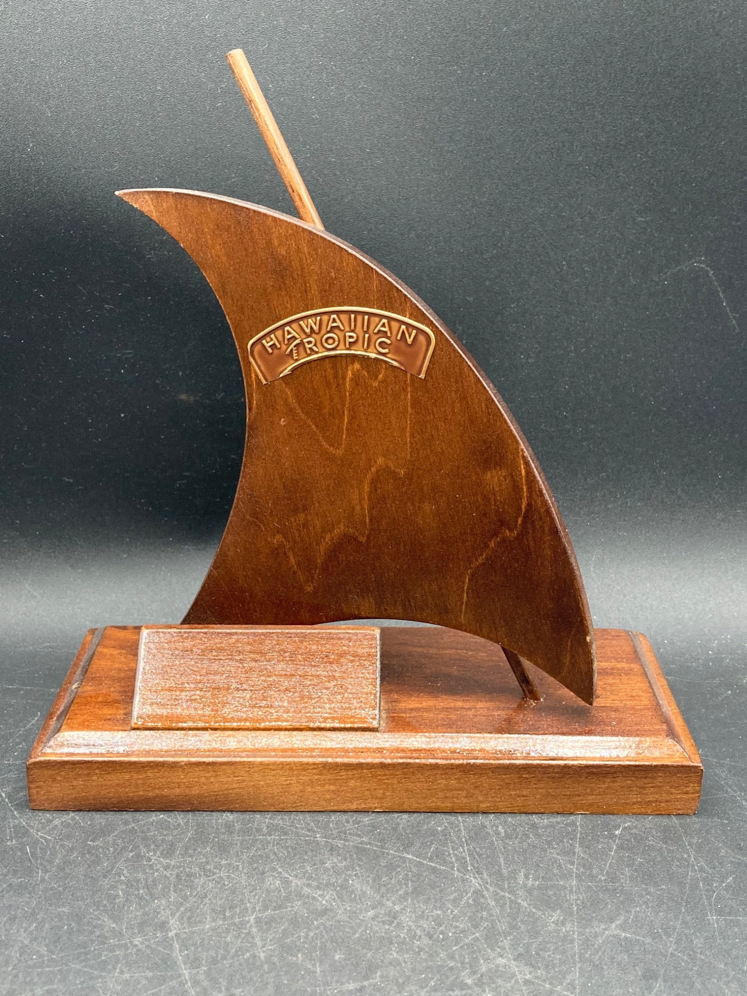 Hawaiian Tropic Wooden Sailboat Award with Display Plaque: This item is a Hawaiian Tropic sailboat-shaped award made of wood. It features a sail design with the 'Hawaiian Tropic' logo fixed to the sail. The award is mounted on a rectangular wooden base that i