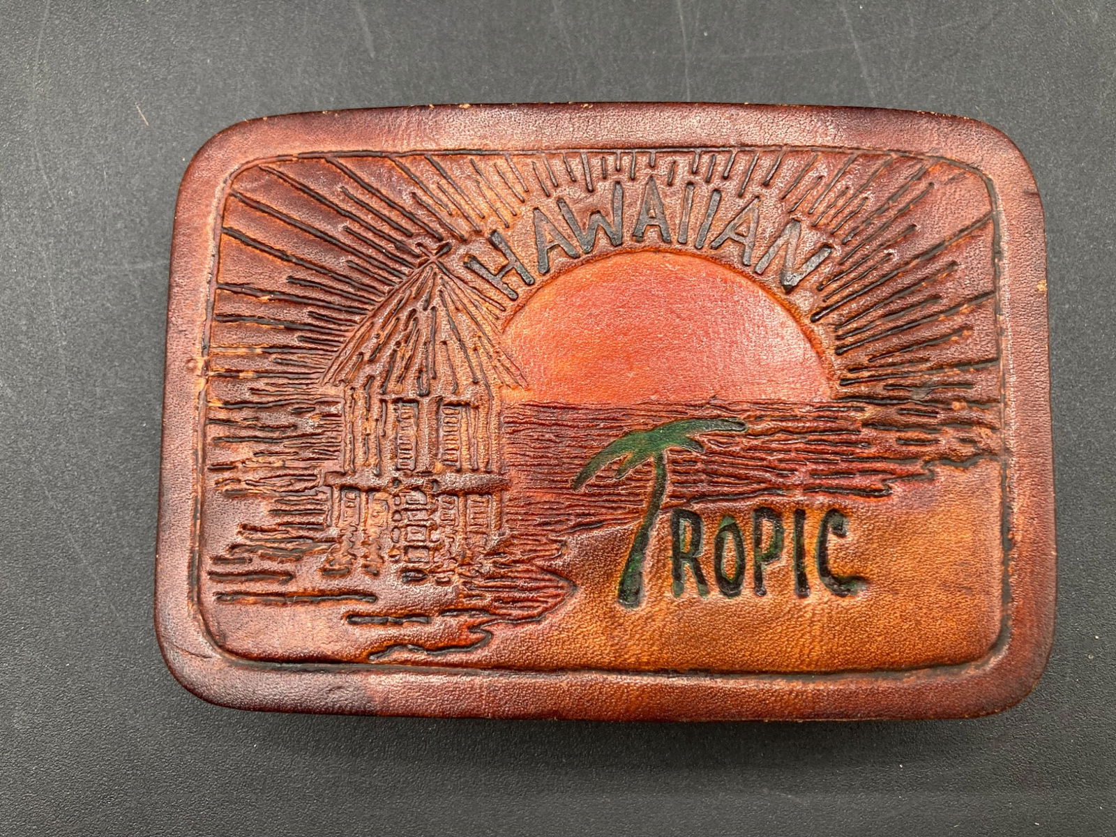 Leather Hawaiian Tropic Belt Buckle with Sun and Hut Design: Rectangular leather belt buckle featuring a Hawaiian Tropic logo with a sun and hut design. The back is metallic, with evident wear and rust. Measures approximately 4 inches long and 2.5 inches wide.