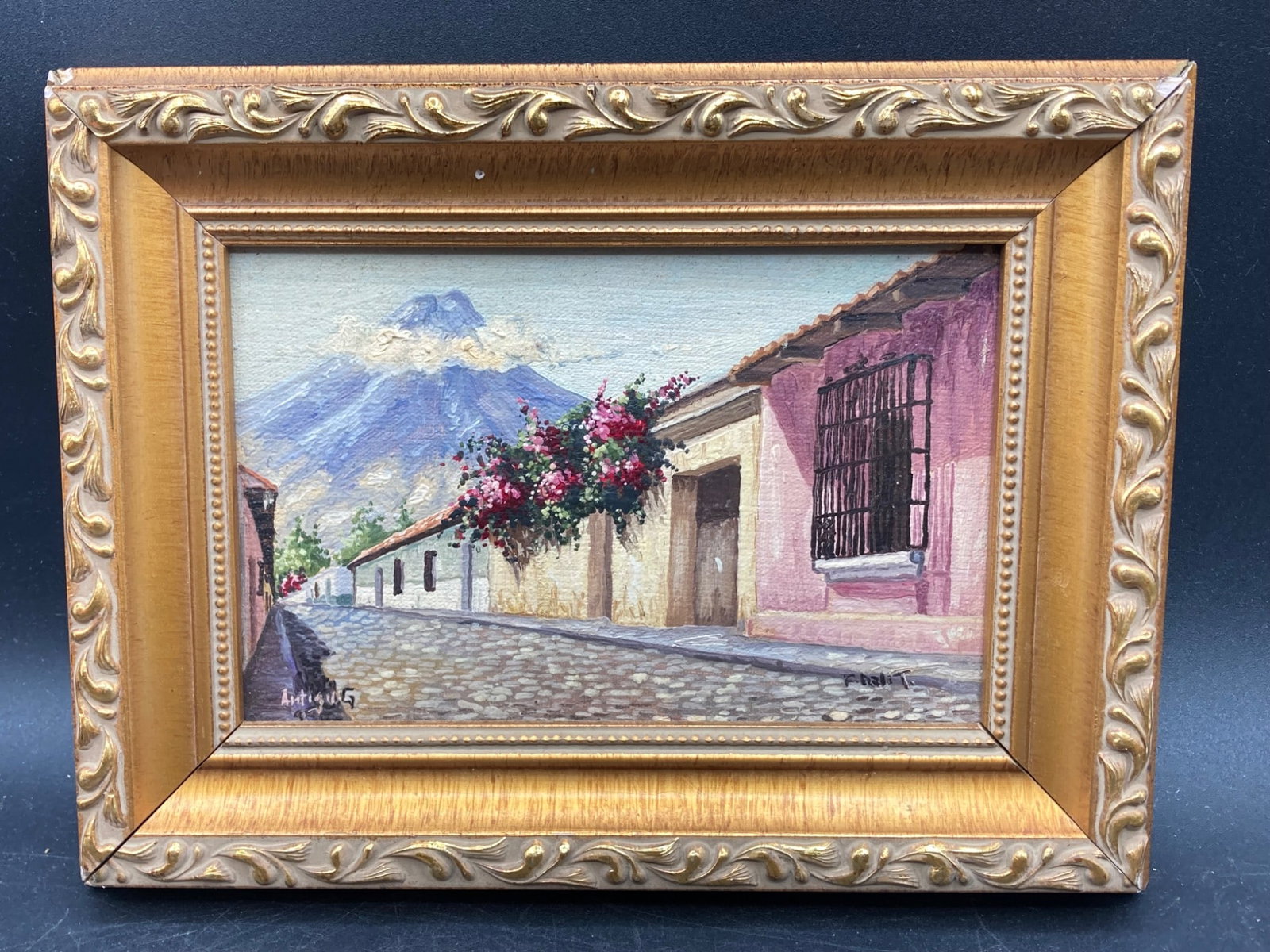 Miniature Antigua, Guatemala Oil Painting: This oil painting on canvas depicts a street scene in Antigua, Guatemala, featuring vibrant bougainvillea flowers and a volcano in the background. The artwork is housed in an ornate gold-colored woode