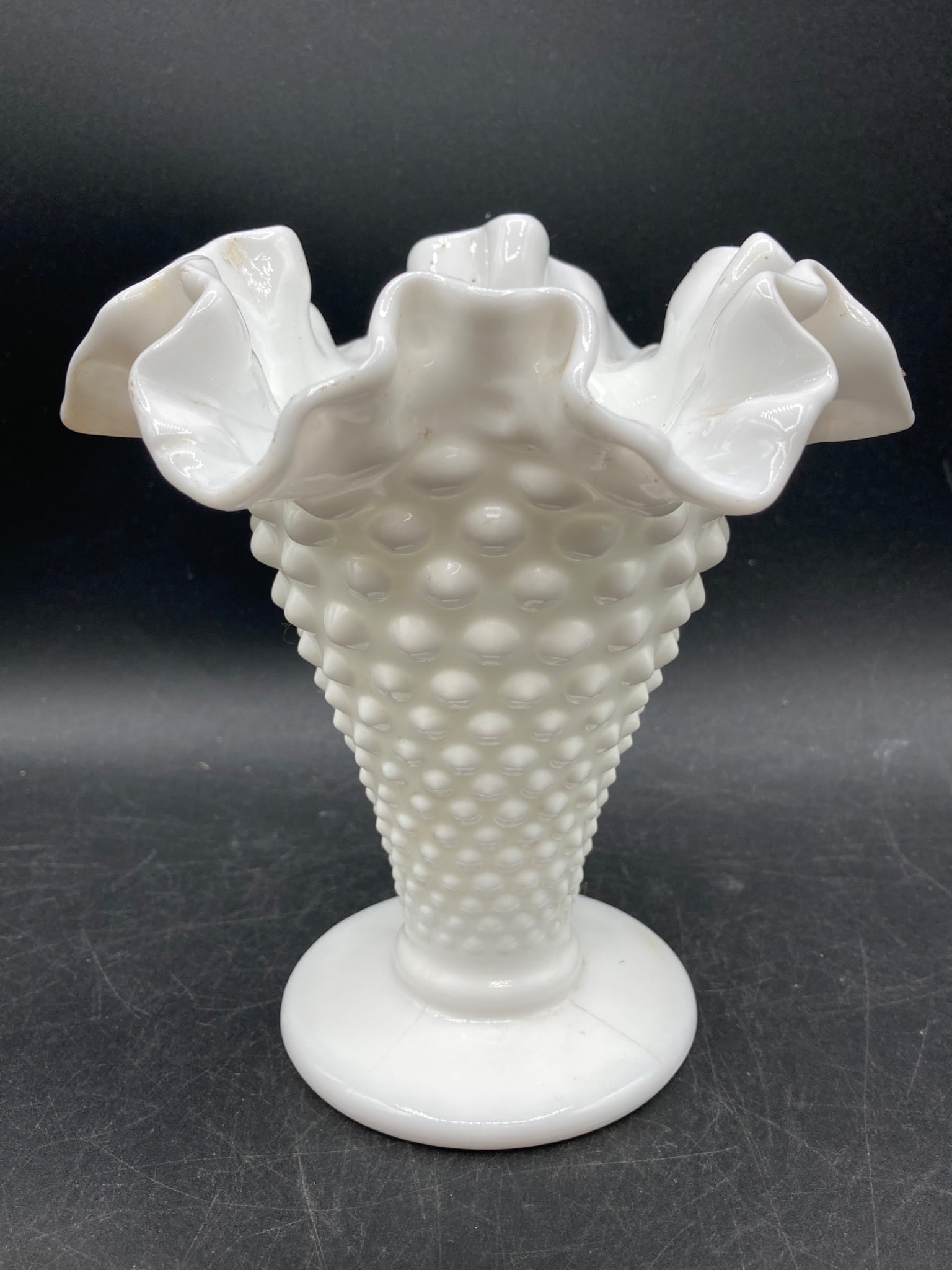 Milk Glass Hobnail Ruffled Vase: This milk glass vase features a hobnail texture and a ruffled edge. The vase stands on a circular flat base, with a tapered body leading to its ruffled rim. It is opaque white with no identified maker