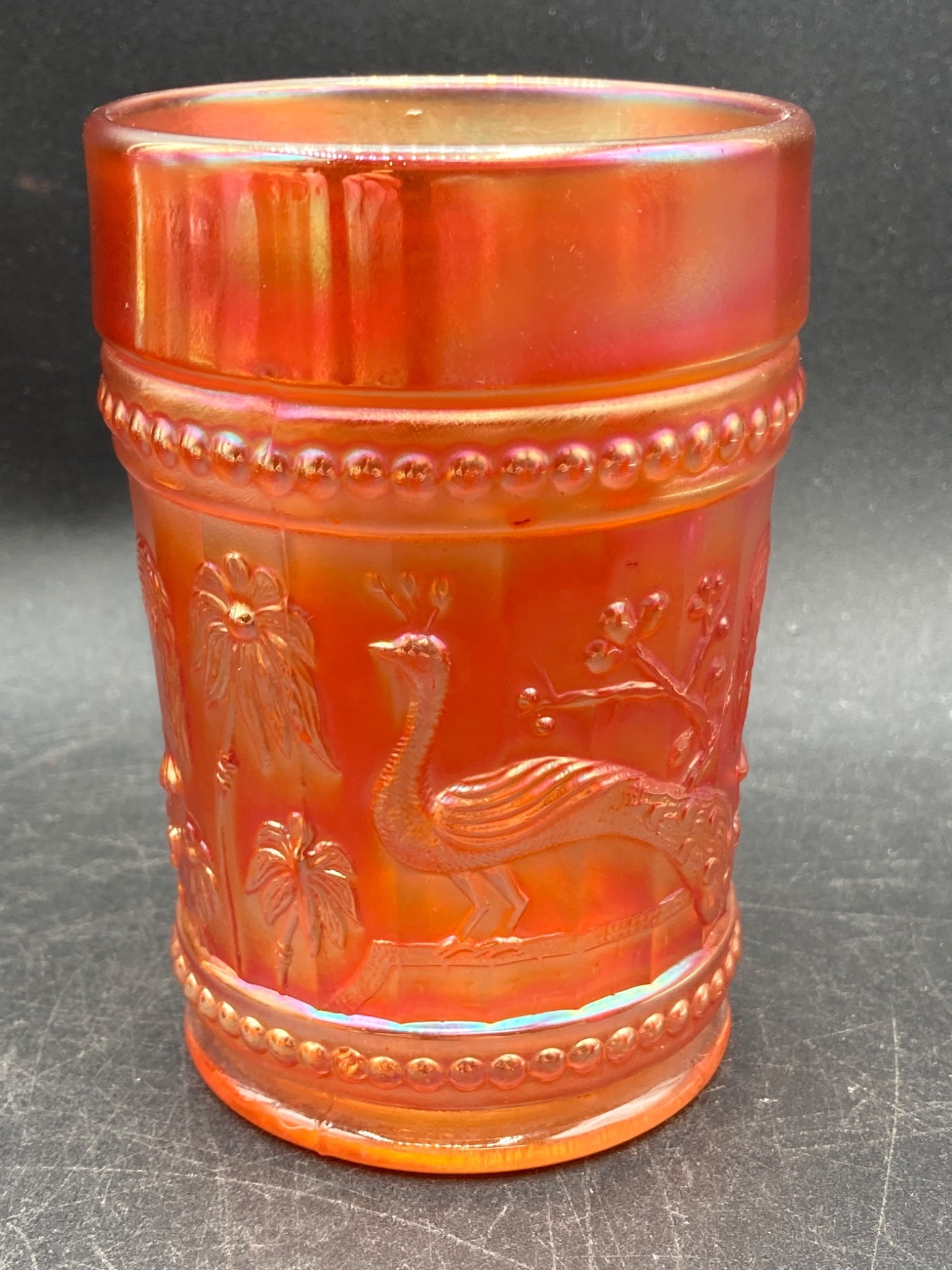 Carnival Glass Marigold Peacock and Palm Tree Tumbler: This is a Carnival glass marigold iridescent tumbler featuring a detailed motif of peacocks and palm trees. The glass has an orange iridescent finish with beaded accents around the body. The glass mea