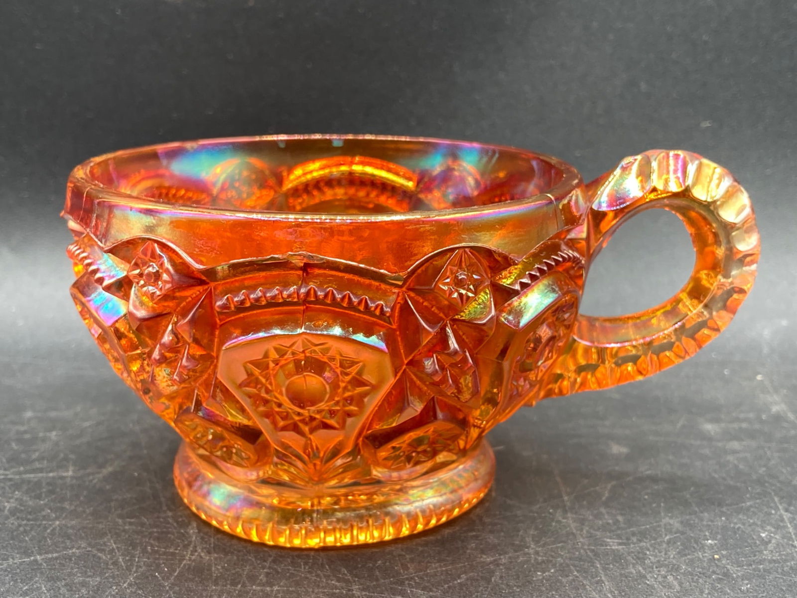 Carnival Glass Marigold Heirloom Tea Cup: This is a Carnival Glass tea cup in a marigold finish featuring a detailed pressed starburst and geometric pattern. The iridescent glass displays a multicolor sheen under light. The base has a matchin