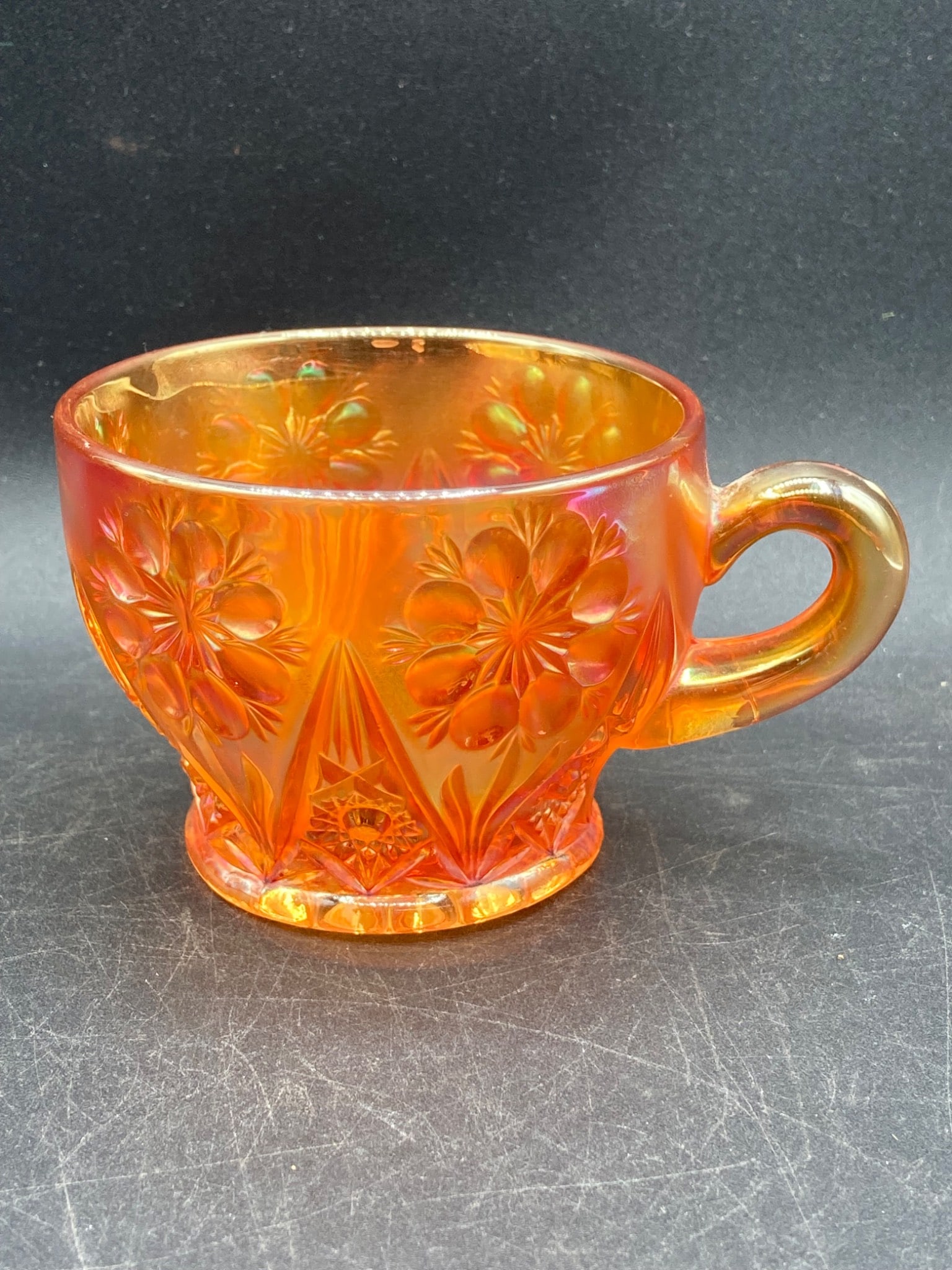 Carnival Glass Marigold Daisies Pattern Tea Cup: This item is a Carnival Glass tea cup with a marigold color and a daisies pattern. The cup features detailed floral motifs and an iridescent finish, typical of carnival glass styles. The base showcase