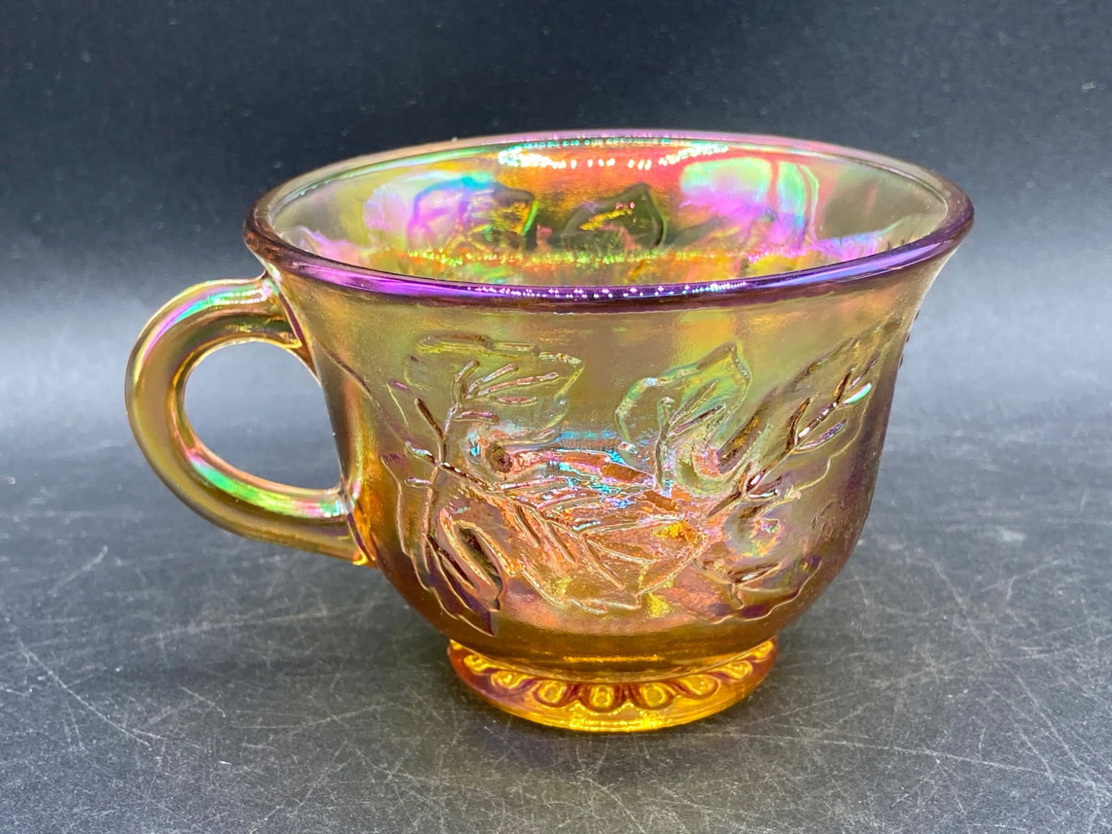 Carnival Glass Amber Grapes and Leaves Tea Cup: This is a Carnival Glass tea cup featuring an iridescent amber finish, with a Grapes and Leaves pattern embossed on the exterior. The base features a decorative floral-like design. The tea cup shows v