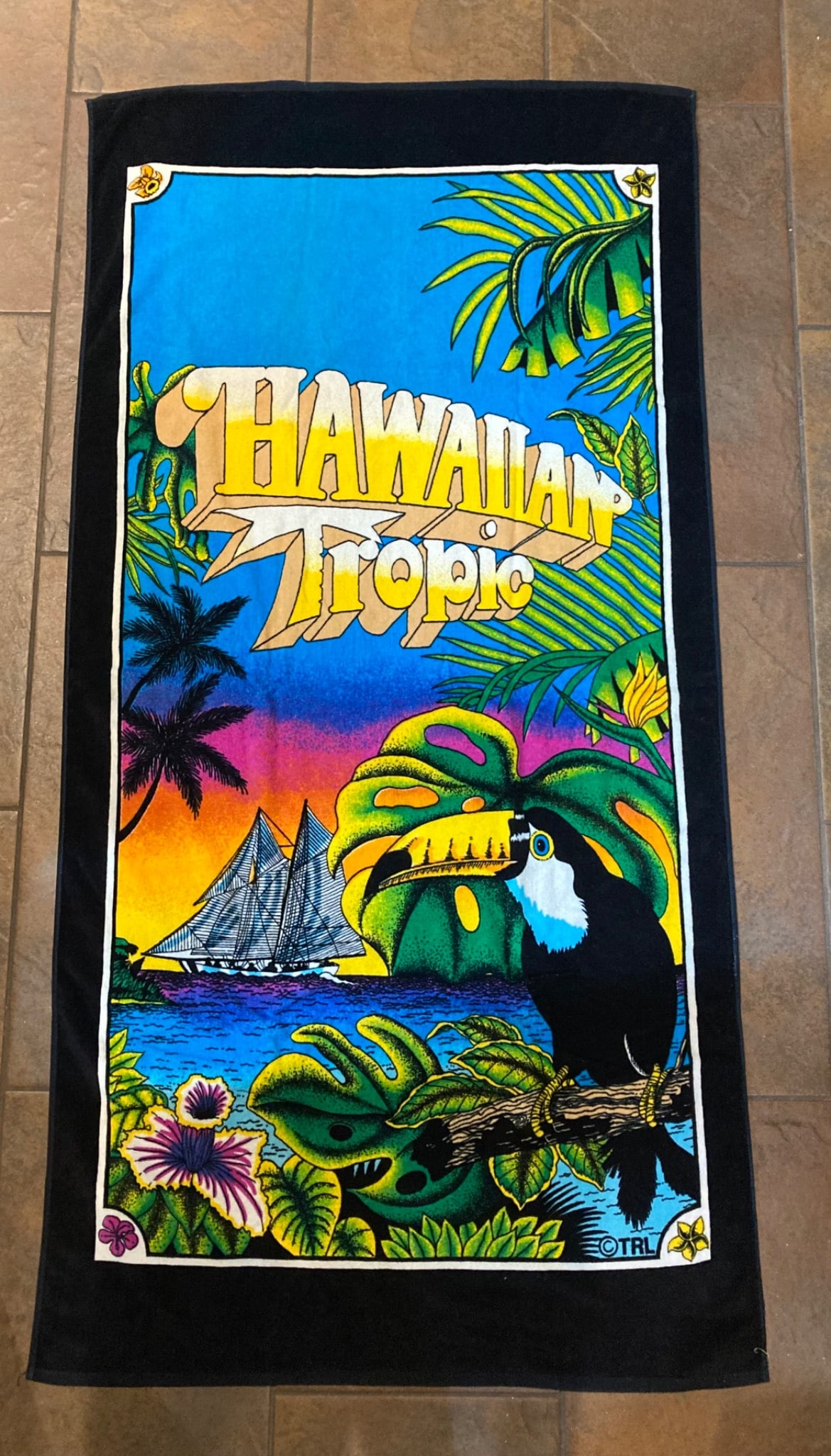 Vintage Hawaiian Tropic Beach Towel: This is a vintage Hawaiian Tropic branded beach towel featuring vibrant tropical imagery, including a toucan, sailboat, and lush foliage against a sunset backdrop. The towel includes the prominent Haw