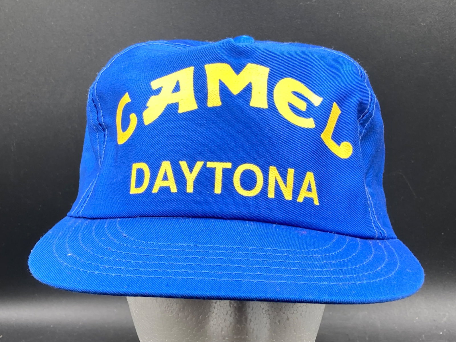 Blue Camel Daytona Snapback Hat, Made in USA: Blue Camel Daytona snapback hat featuring yellow text embroidery for Camel and Daytona on the front. Manufactured by ROMAN CAPS in the USA, the hat is made from 100% cotton and has an adjustable plast