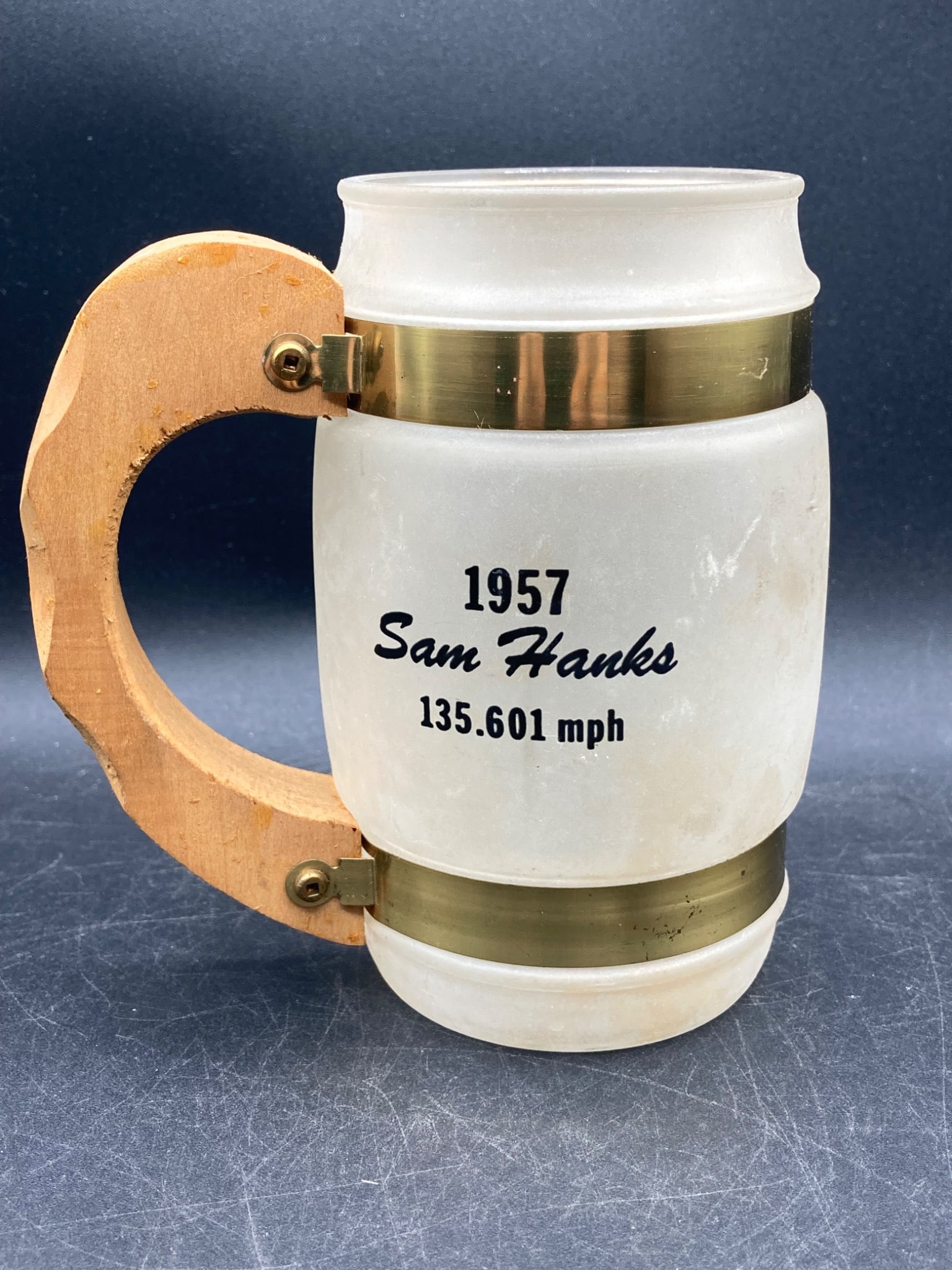 Siesta Ware 1957 Indy 500 Winner Sam Hanks Mug: Frosted glass mug produced by Siesta Ware, commemorating the 1957 Indianapolis 500 win by Sam Hanks driving the Belond Exhaust Special, with a speed of 135.601 mph. Features a wooden handle attached w