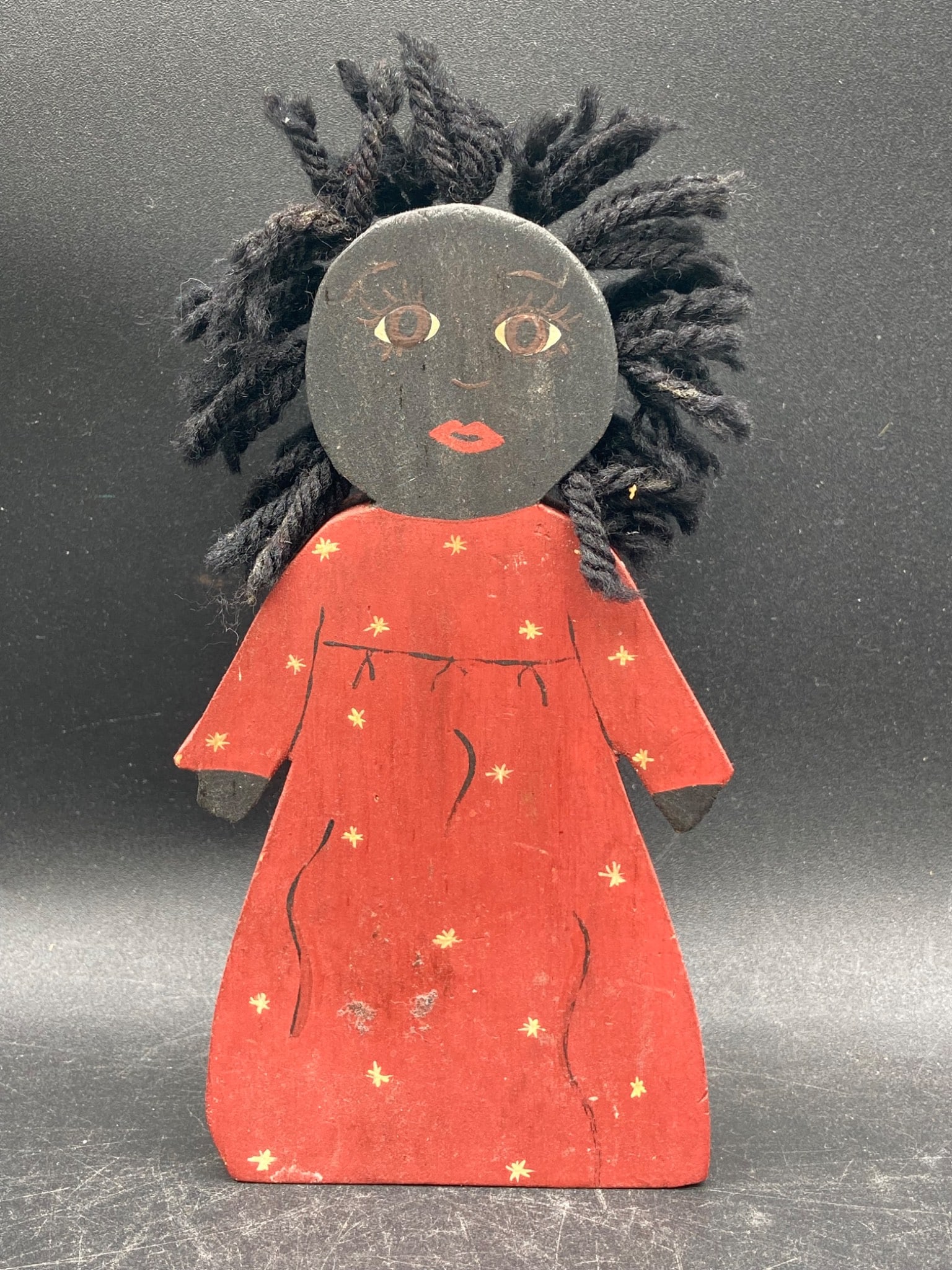 Black Americana Wooden Figure: Wooden figure featuring hand-painted details, depicting a face and a body clad in a red dress adorned with yellow star accents. The figure measures approximately 6 inches in height. Signature visible 