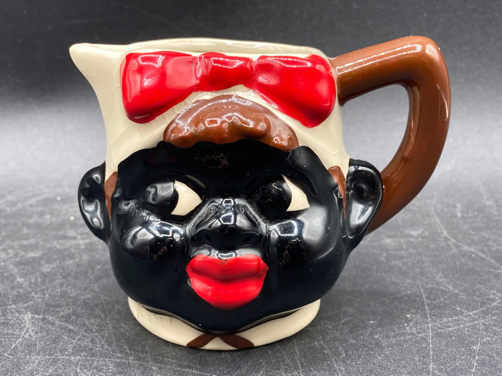 Back Americana Porcelain Creamer with Red Bow and Brown Handle: This is a porcelain creamer noted for its back Americana style depicting a figure with a red bow and a brown handle. The approximate height is 3 inches. The creamer's exterior shows age-related crazin