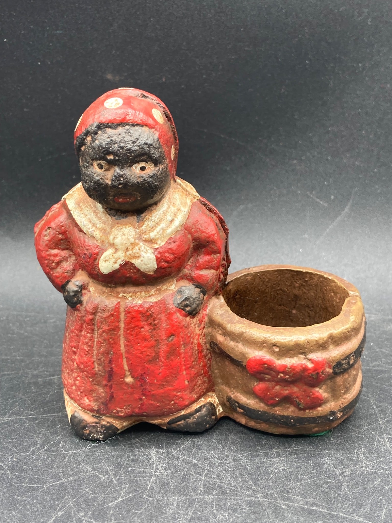 Antique Black Americana Cast Iron Coin Holder: This is an antique Black Americana cast iron coin holder with a hand painted design. The piece depicts a figure dressed in a polka-dot red headscarf and dress, standing next to a barrel. The item meas