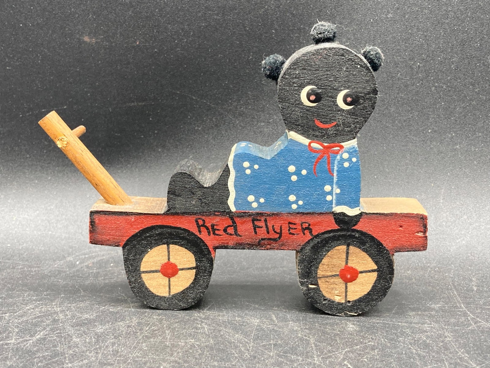 Black Americana Painted Wood Red Flyer & Girl Figure: This item is a handcrafted wooden Red Flyer wagon with a fixed girl figure. Measuring approximately 5 inches in length, the figure features painted details, including a blue dress with white dots, a r