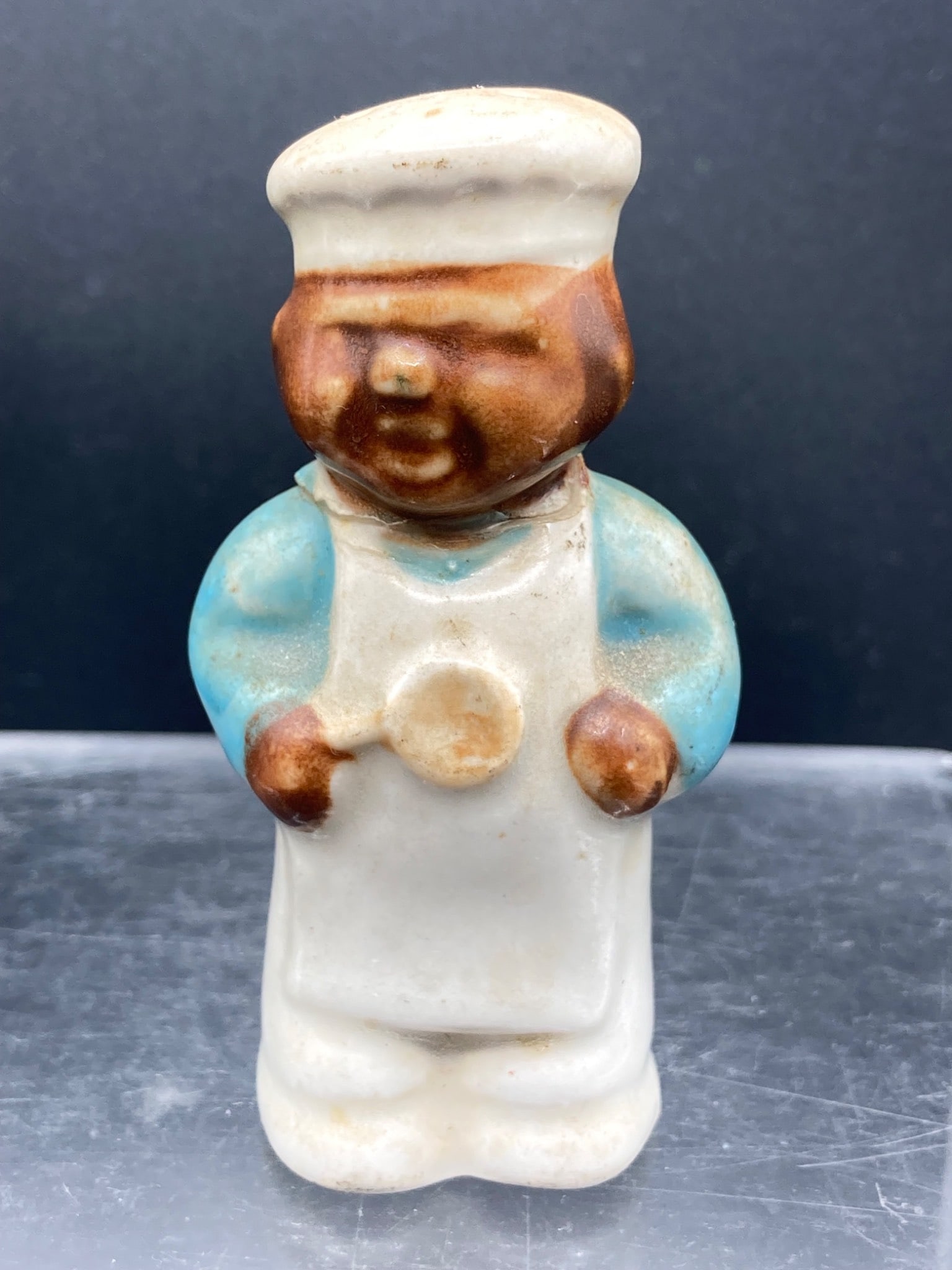 Black Americana Porcelain Baker Pepper Shaker: This porcelain pepper shaker depicts a baker wearing a white chef’s hat, apron, and blue shirt, with hand-painted details. It is crafted in the form of Black Americana artwork. The shaker features h