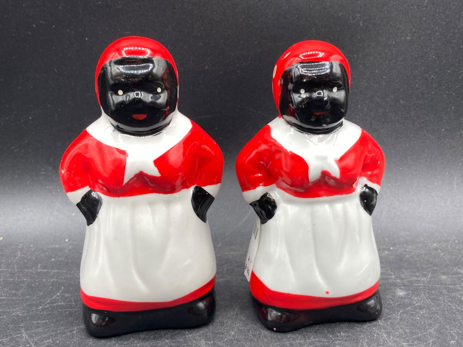 Black Americana Porcelain Salt & Pepper Shaker Set: This lot includes a pair of porcelain salt and pepper shakers, approximately 4 inches tall each, depicting characters dressed in red and white attire. The set features plastic stoppers at the base for
