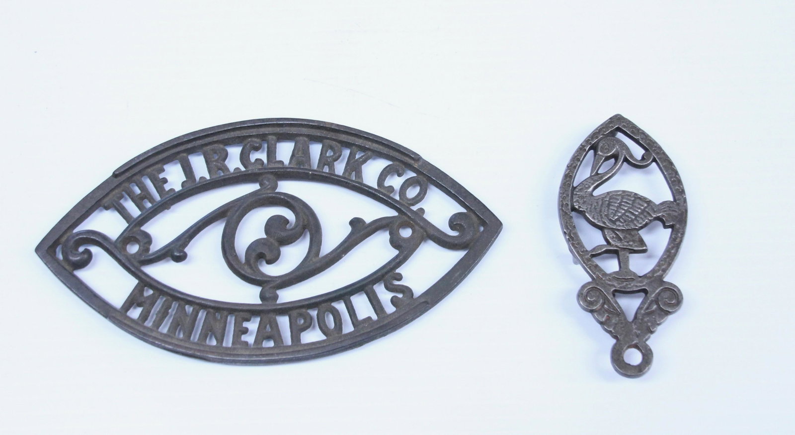 PAIR OF CAST IRON TRIVETS: SMALL TRIVET WITH BIRD MOTIF AND ADVERTISING TRIVET (THE JR CLARK CO. MINNEAPOLIS) Dimensions: 4" TO 7" L