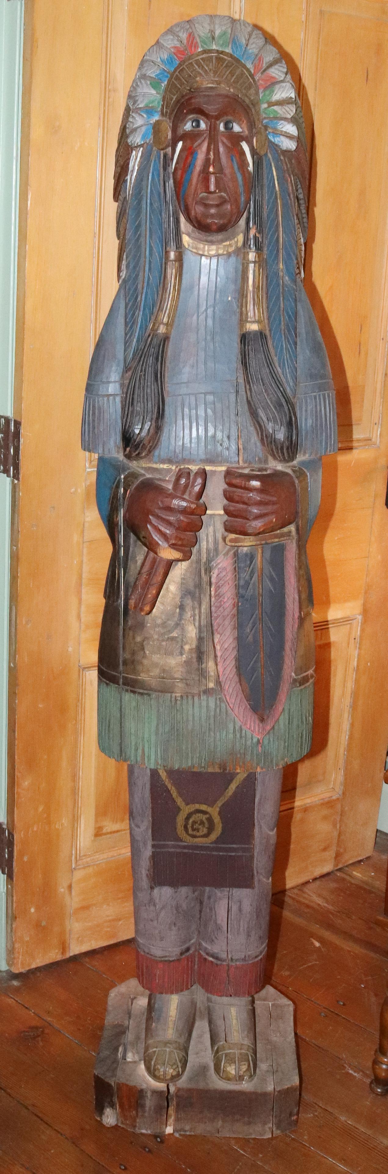 WOODEN CIGAR STORE INDIAN: 67" H NATIVE AMERICAN WITH LONG FEATHERED HEADDRESS HOLDING A GROUP OF CIGARS, PAINTED Size: 67"x 13 1/2"x 16" Terms: PLEASE READ THIS CAREFULLY AND FULLY BEFORE BIDDING - THIS IS AN ON-SITE AND LIVE