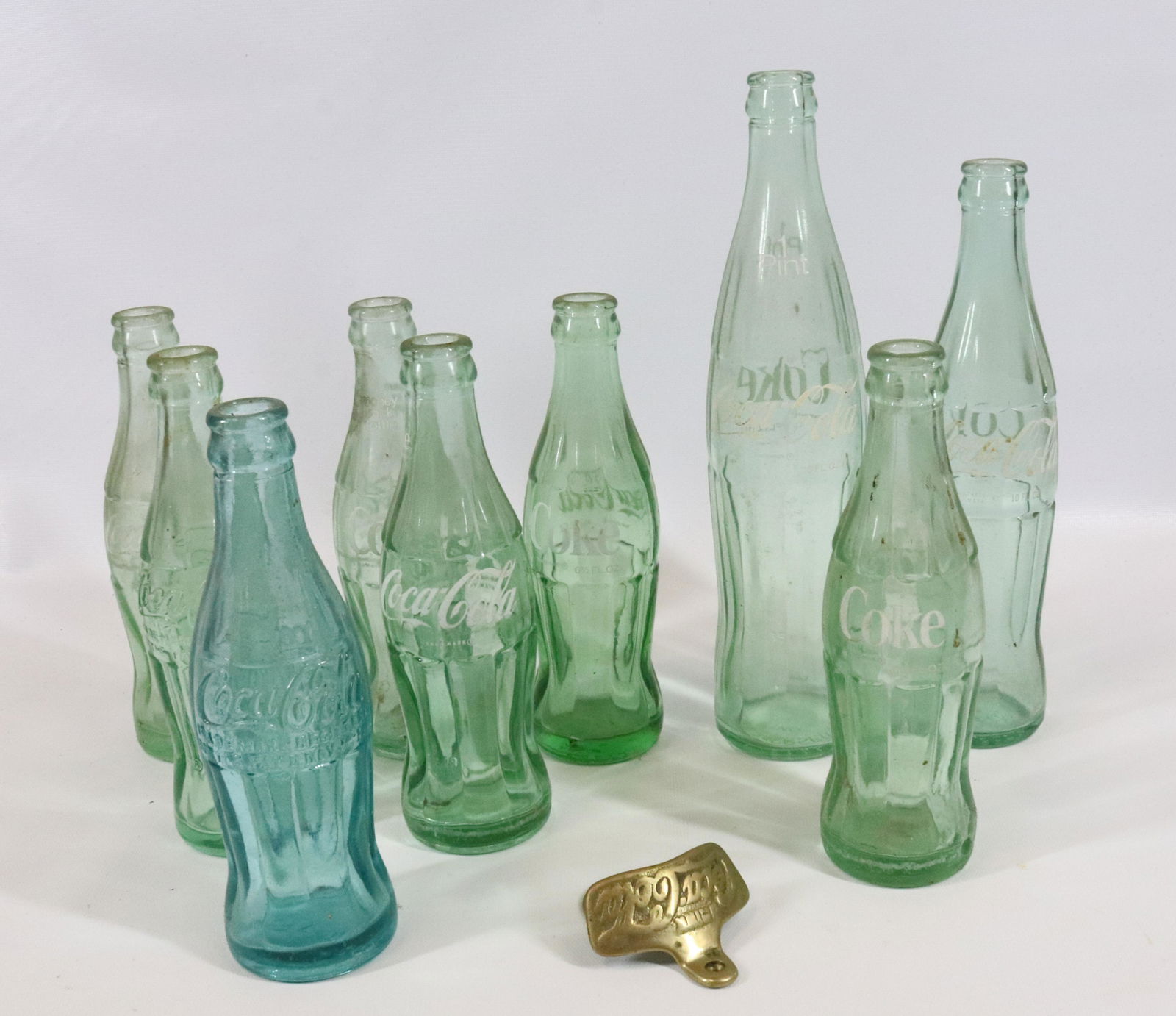 LOT OF COCA COLA BOTTLES AND OPENER: NINE AQUA COLORED BOTTLES AND BRASS BOTTLE OPENER. LARGEST BOTTLE 11 1/4" H.
