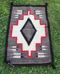 NAVAJO RUG IN BLACK, GRAY/BROWN, RED, CREAM WITH CENTRAL MOTIF SURROUNDED BY FEATHERS.  33 1/2" X 21