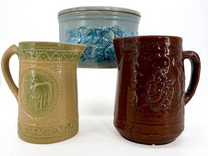 LOT OF COUNTRY POTTERY PITCHERS, CROCK: BLUE EMBOSSED CAKE CROCK WITH LID (GRAPE/LEAF DECORATION). 6" X 9 1/2" D. YELLOW WARE EMBOSSED MILK PITCHER IN GREEN/YELLOW WITH COWS 7 3/8" H. AND BROWN EMBOSSED PITCHER WITH GRAPE PATTERN 7 7/8" H.