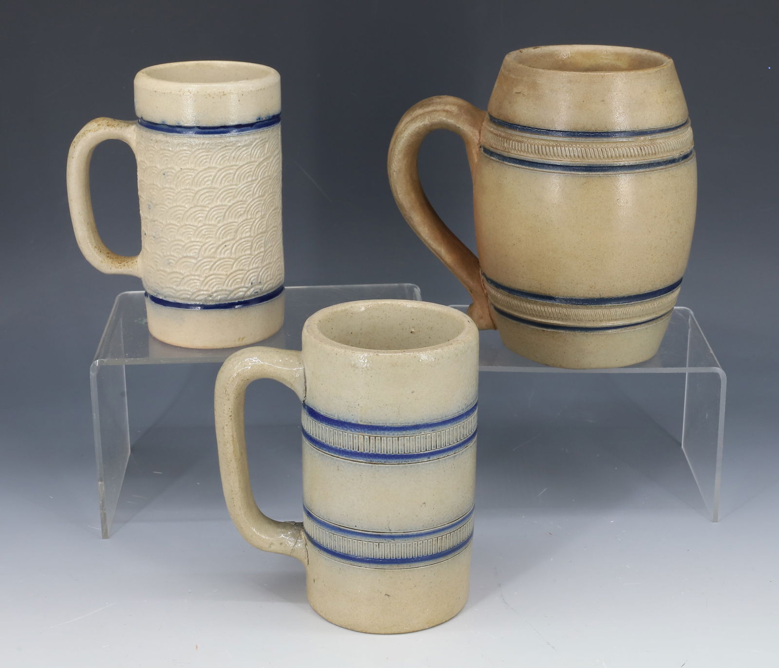(3) STONEWARE BEER MUGS: VARIOUS MOLDED STONEWARE MUGS WITH BLUE BANDING. LARGEST HAS SHAPELY APPLIED HANDLE. MRKD. NO. 19 ON BOTTOM. 5 1/2" H. PLEASE READ CAREFULLY - Terms for Purchasing and Shipping of Auction Items. Pleas