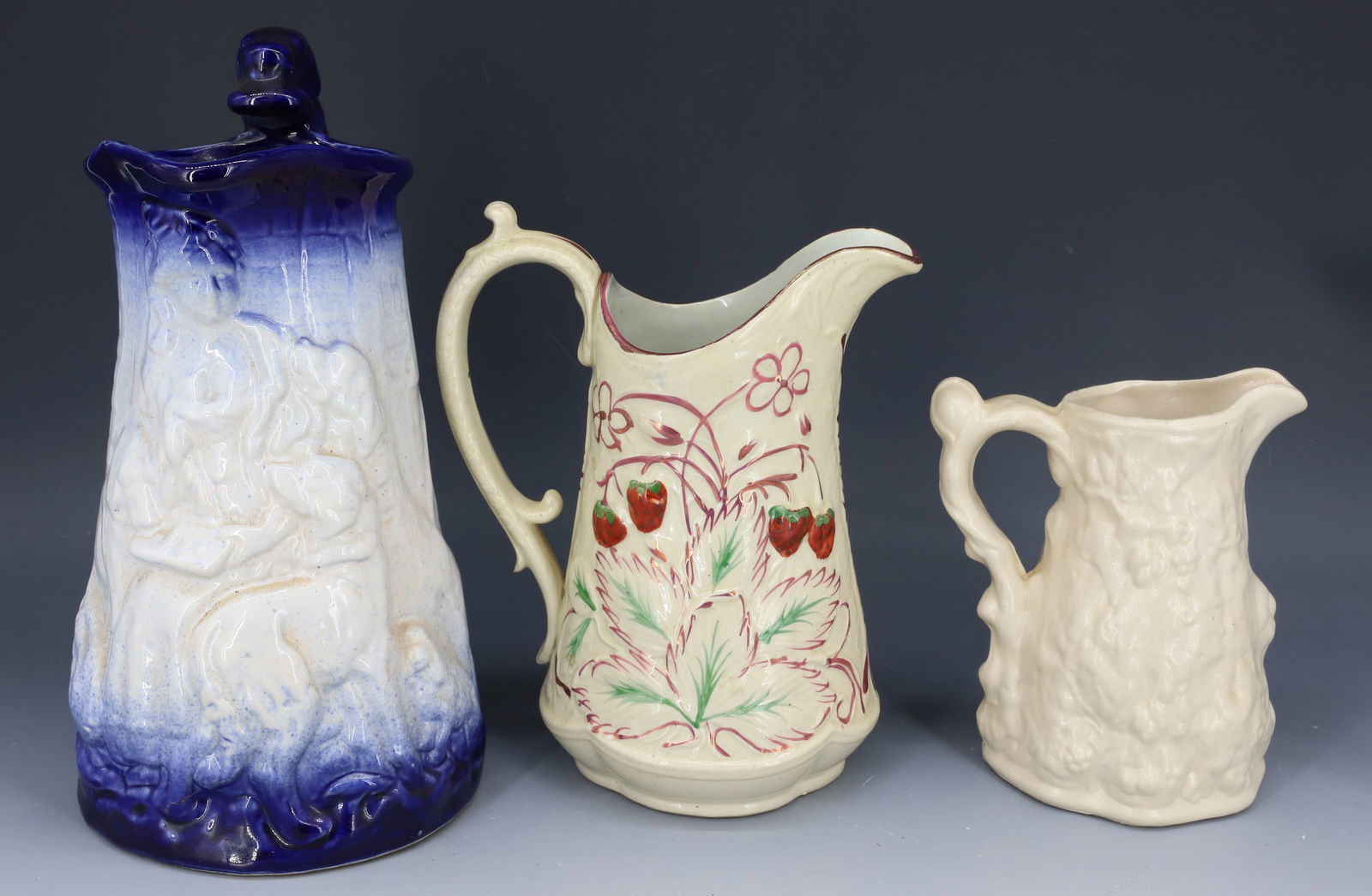(3) EMBOSSED POTTERY PITCHERS: SMALLEST HAS ALL-OVER EMBOSSED DECORATION 5 1/2" H., STRAWBERRY PATTERN PITCHER WITH LUSTER HIGHLIGHTS AND LARGE EMBOSSED BLUE/WHITE PITCHER WITH DOG HANDLE 10" H. ALL UNMARKED. PLEASE READ CAREFULLY