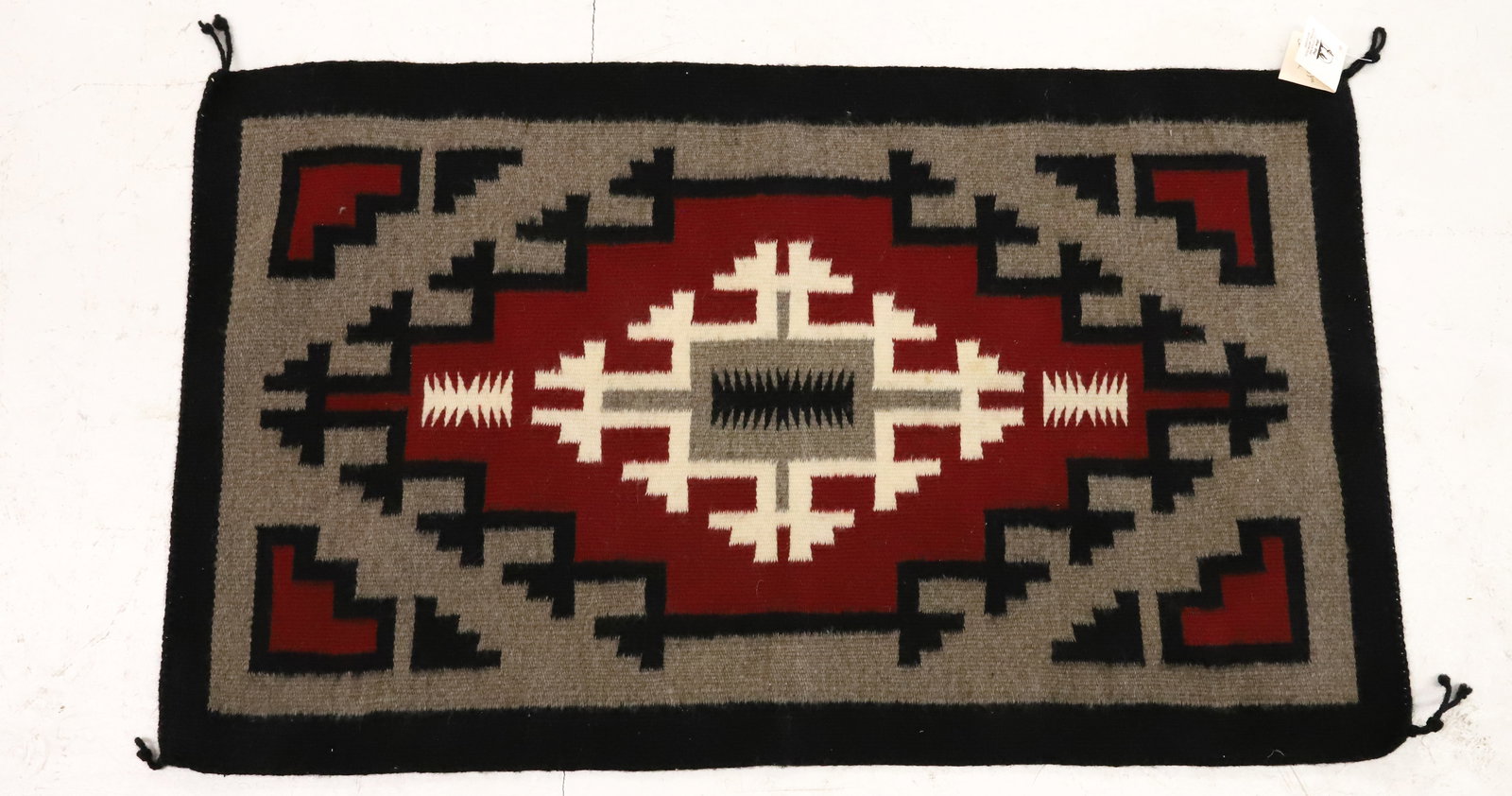 VINTAGE NAVAJO RUG W/ TAG: BLACK / GRAY/ RED / CREAM. TAG "GREY WOLF, MADE BY PRICELLA YAZZIE". 36" X 21". PLEASE READ CAREFULLY - ITEMS MUST BE PAID FOR THROUGH LIVE AUCTIONEERS OR WITH CASH OR CHECK IN PERSON - Terms
