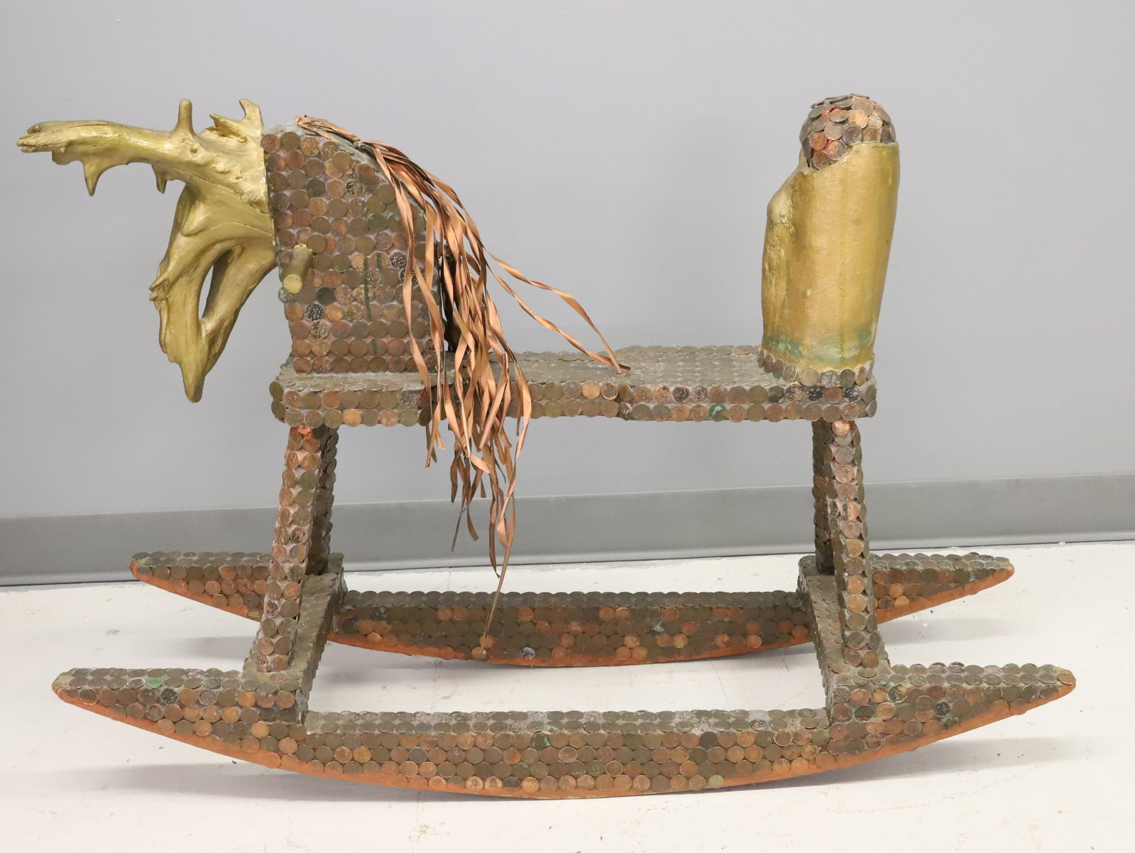 FOLK ART ROCKING HORSE: WOODEN ROCKING HORSE COVERED IN PENNIES. COPPER COLORED RIBBON MANE AND ABSTRACT WOODEN "ANTLER" HEAD. 40 1/2" L. PLEASE READ CAREFULLY - ITEMS MUST BE PAID FOR THROUGH LIVE AUCTIONEERS OR WITH CASH