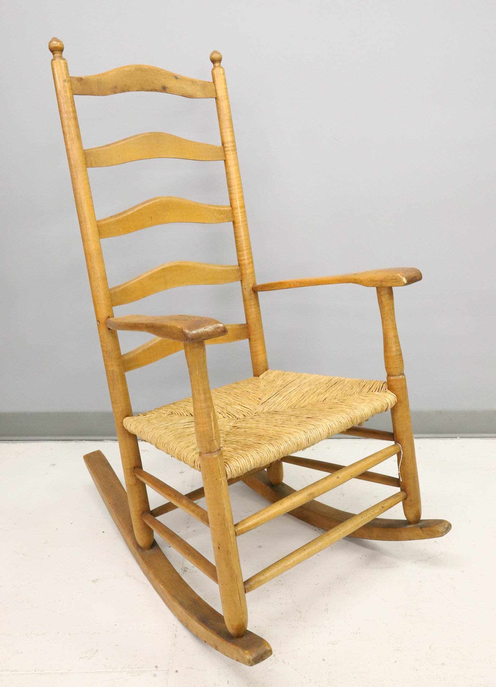19TH C. MAPLE ROCKING CHAIR: TIGER MAPLE WITH LADDER BACK. RUSH SEAT. PEGGED CONSTRUCTION. 45" X 23 1/2" X 17". PLEASE READ CAREFULLY - ITEMS MUST BE PAID FOR THROUGH LIVE AUCTIONEERS OR WITH CASH OR CHECK IN PERSON - Terms for