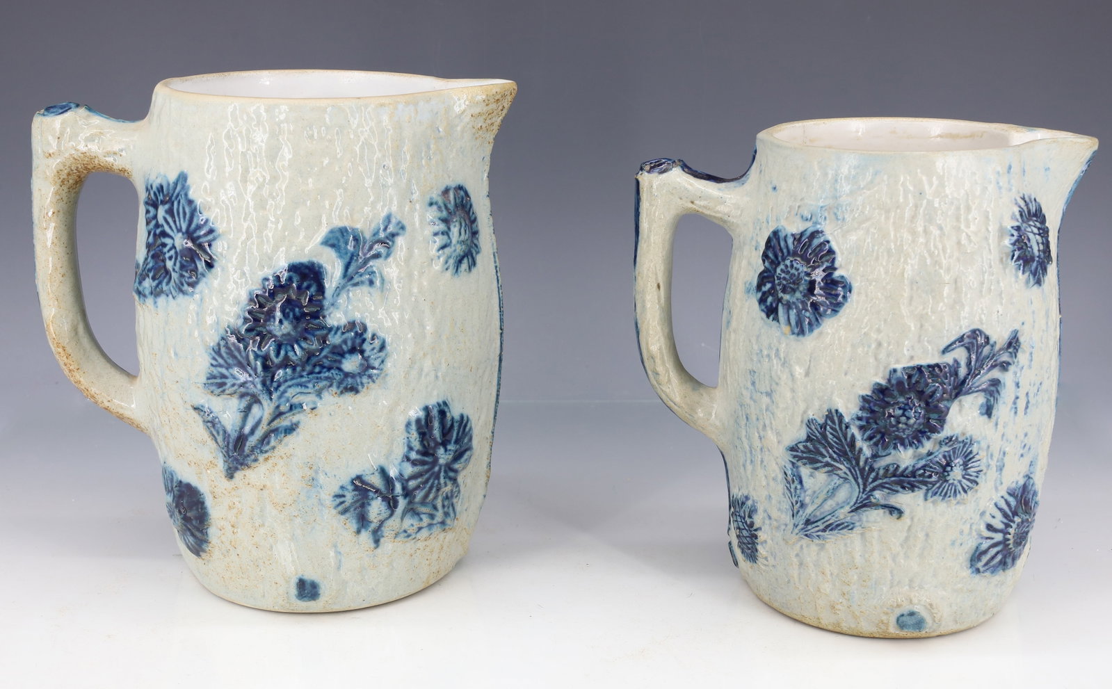 (2) WHITE'S UTICA STONEWARE PITCHERS: MOLDED PITCHERS WITH BARK SURFACE AND BLUE FLORAL DESIGN - ONE MARKED 12 AND ONE MARKED 18 ON BOTTOM - 6 3/4" AND 7 1/4" H PLEASE READ CAREFULLY - ITEMS MUST BE PAID FOR THROUGH LIVE AUCTIONEERS OR WI