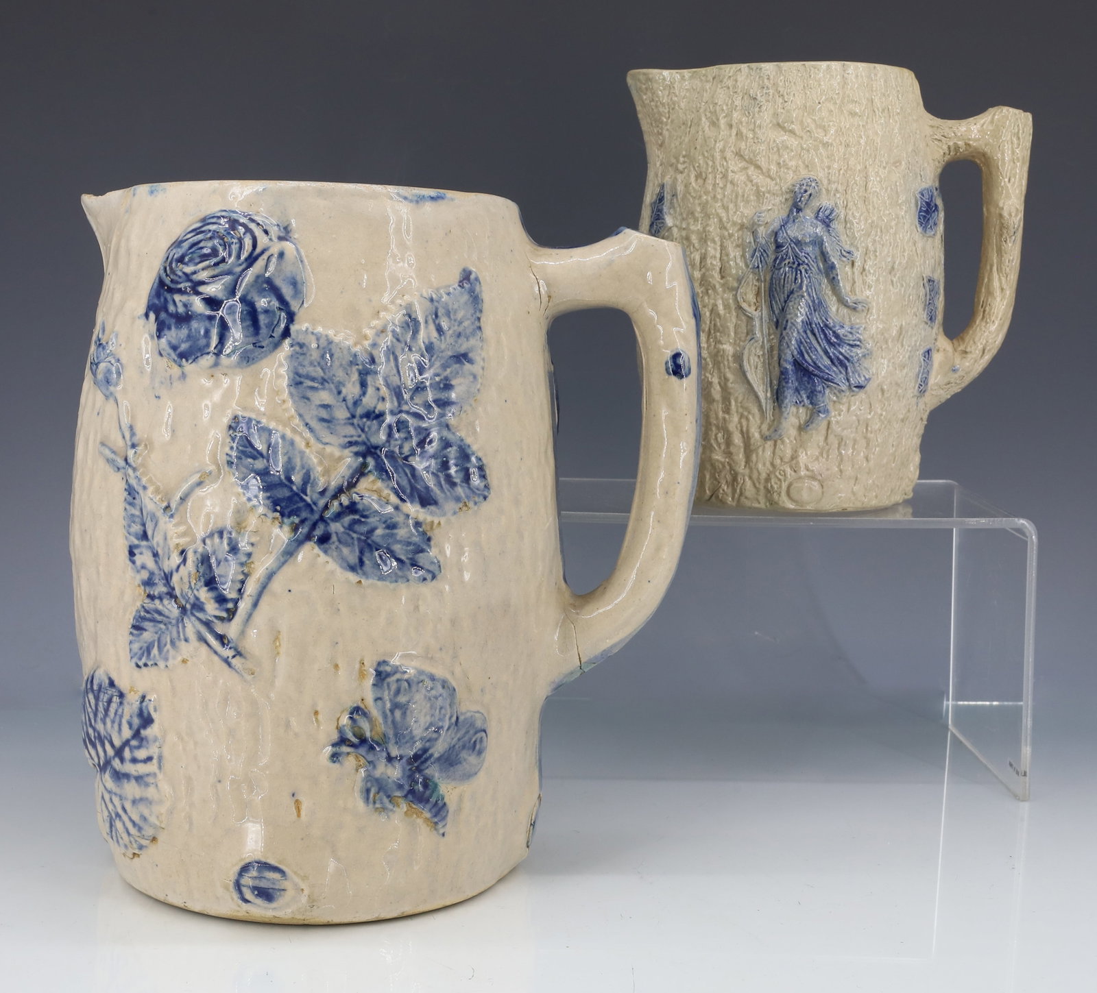 (2) WHITE'S UTICA STONEWARE PITCHERS: MOLDED PITCHERS WITH BLUE HIGHLIGHTS. SMALLER DEPICTS HUNTRESS WITH BOW/ARROWS. 6 1/8" H. LARGER DEPICTS LEANING MAN AND FLORAL DECORATION. 8" H. (IMPRESSED 6 ON BOTTOM). PLEASE READ CAREFULLY - ITEMS