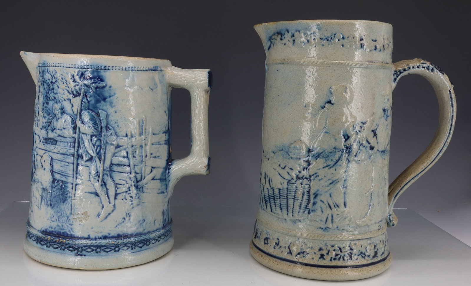 (2) WHITE'S UTICA STONEWARE PITCHERS: MOLDED PITCHERS WITH BLUE HIGHLIGHTS. SMALLER HAS MOLDED SCENE ON EITHER SIDE (INTERIOR AND EXTERIOR SCENES) 7 5/8" H. (MRKD. 10 ON BOTTOM) TALLER HAS SCENES OF MEN AND WOMEN ON BOTH SIDES. MRKD. 5 ON