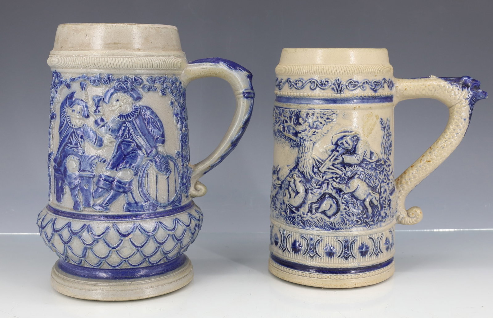 (2) WHITE'S UTICA STONEWARE MUGS, STEINS: LARGER HAS TWO MEN DRINKING ASIDE OF A KEG WITH VERSE ON BACK. COBALT BLUE DECORATION. MRKD. 38 ON BOTTOM. 7 1/4" H. OTHER, HAS HUNTING SCENE AND VERSE ON BACK. MRKD. 45. 6 3/4" H. PLEASE READ CAREFUL