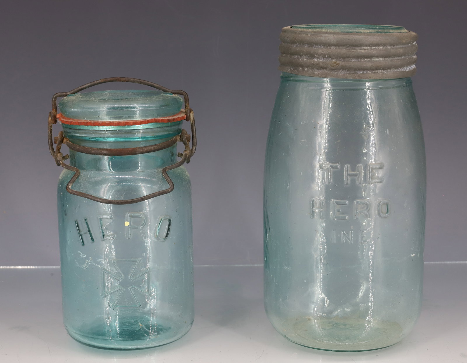 (2) "HERO" FRUIT JARS: AQUA JARS INCLUDING: "THE HERO INE " JAR WITH PLAIN GLASS INSERT LID. BOTTOM MRKD. "69, PATD NOV 26, 1867" 7 1/4" TOTAL HEIGHT. WITH SMALLER "HERO" WITH CROSS. BOTTOM MRKD. 29. WIRE CLOSURE W/ LID
