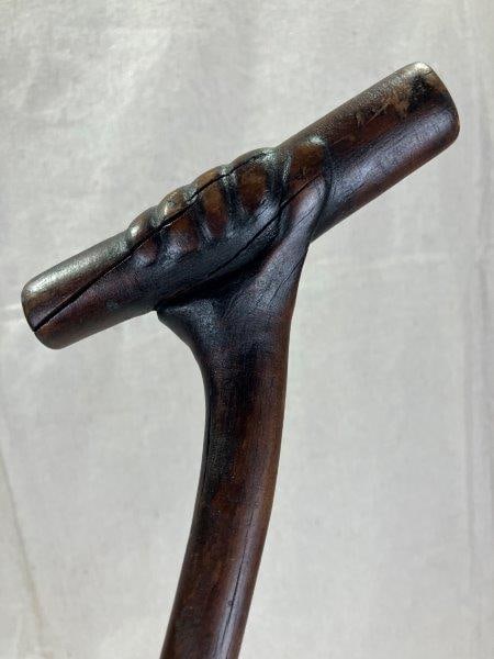 Folk Art Carved Cane: This distinctive folk art cane features a carved hand grasping the handle, knotty wood shaft adds rustic charm, while a brass cap at the base provides a refined finishing touch. Measures approx. 37 5/