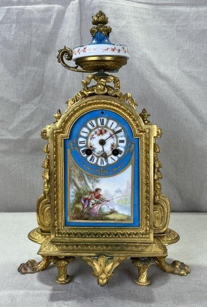 Miroy-Requier & Co. Gilt Bronze Mantel Clock: Elegant gilt bronze mantel clock featuring a richly detailed pastoral scene on the clock face, depicting a pair fishing, framed by a gold border. Topped with a painted glass urn and foliate finial, an