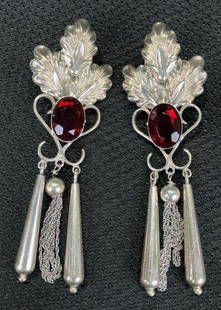 Sterling Silver Ruby Toned Stone Dangle Earrings: A striking pair of sterling silver dangle earrings, each featuring a central ruby-toned stone framed by three-dimensional leaf motifs and intricate scrollwork. The design is accented with a central ha