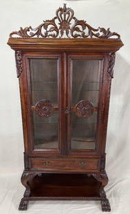 Ornately Carved Mahogany Curio Cabinet: A grand and richly detailed curio cabinet featuring an ornate carved crest, dark mahogany finish, and central medallions adorning the paneled doors. The dramatic form and deep tone make it a standout