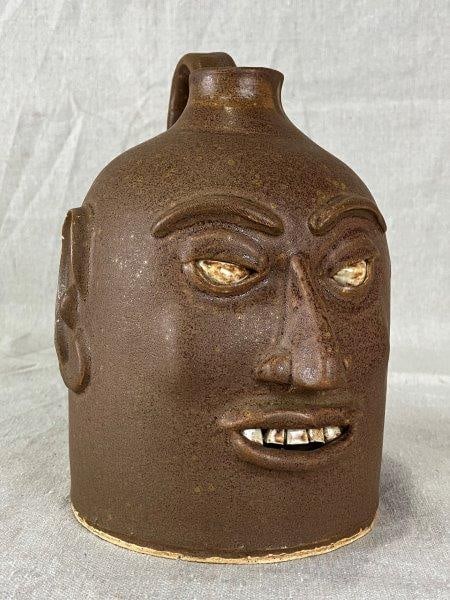 Southern Stoneware Face Jug: Features a warm brown glaze, applied facial features with glossy glazed eyes and teeth, and an applied handle. Inscribed on base "Kevin L. Smith 04 Mud Baby Columbia, SC." Measures approximately 7 5/8