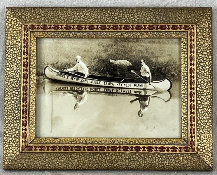 Early 20th Century Canoeing Photograph: Striking black-and-white photograph depicting two men in a canoe adorned with the names of prominent U.S. towns and a nice reflection in the water. Measures approximately 7 1/8" H x 9 1/8" W x 1/2" D.