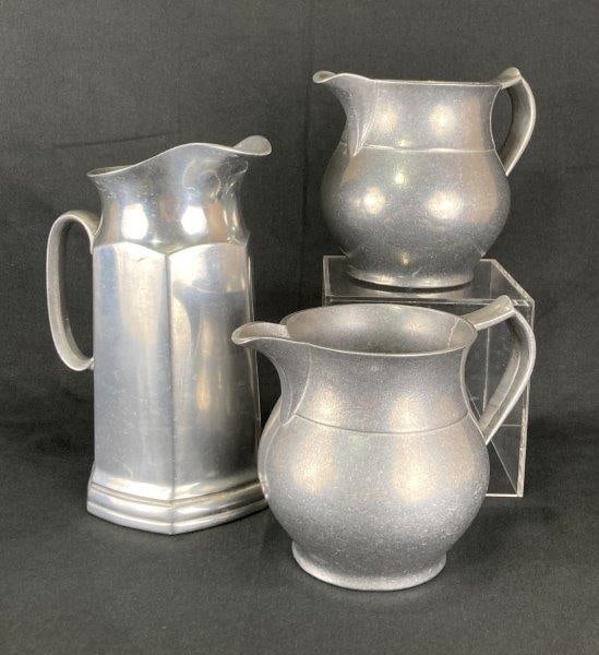 Three Pewter Pitchers: Group of three pewter pitchers, each showcasing distinctive forms characteristic of traditional pewter craftsmanship. The tallest features an elongated body with hexagonal sides, while the two smaller