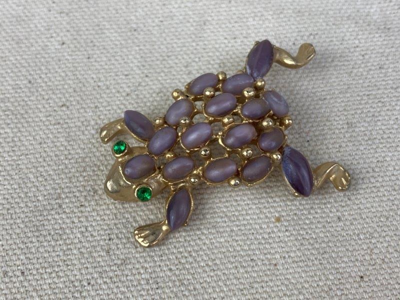Gold-Tone Frog Brooch: Whimsical gold-tone frog brooch set with purple cabochon stones across its body and accented by green gemstone eyes. Unmarked. Measures approximately 1 3/4" long and 1 1/4" wide. In good condition.