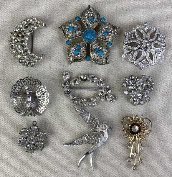 Lot of Nine Costume Jewelry Brooches Including Monet, Sarah Coventry, and Swarovski: A lot of nine costume jewelry brooches in silvertone and goldtone finishes, accented with rhinestones and turquoise-style stones. Makers represented include Monet, Sarah Coventry, and Swarovski. The b