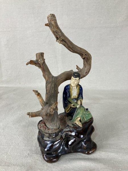 Chinese Mudman Scholar Sculpture: Fine sculpture depicting a scholar seated in a thoughtful pose beneath a carved wooden tree, combining natural materials with refined craftsmanship. Measures approximately 11 1/4" H x 6 1/4" W x 5" D,