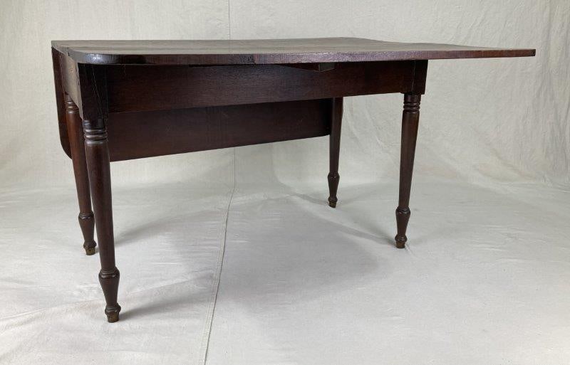 Walnut Drop Leaf Table: Drop-leaf table with two hinged leaves and finely turned legs, exhibiting a warm aged patina. With the leaves raised, it measures approximately 27" H x 48 5/8" W x 41 7/8" D. Shows shrinkage to the to