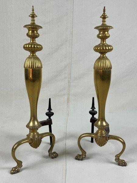 Ornate Brass Andirons: Early 20th century brass andirons featuring elegant curving forms and decorative beading around the base, raised on stylized paw feet. The set retains its original iron log stocks, which have been cut