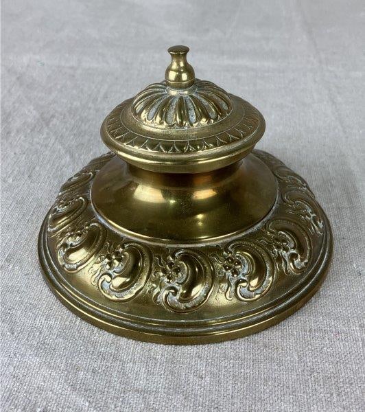 Brass Inkwell: Finely cast brass inkwell featuring an ornate floral motif and warm, lustrous metal tones throughout, exemplifying decorative craftsmanship. Measures approximately 3 3/8" H x 4 7/8" in diameter. Shows