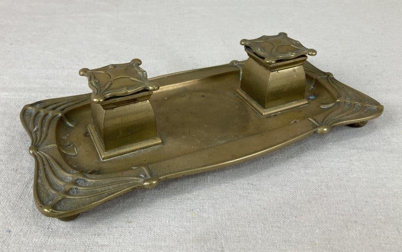 Brass Inkwell: Elegant brass inkwell featuring raised Art Nouveau-style decoration on the rim and twin inkwell lids, set upon a footed tray base. Exhibits a warm aged patina. Measures approximately 2 3/8'' H x 11''