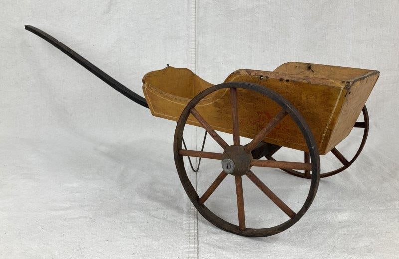Vintage Wheelbarrow Toy: Wooden pull-toy wheelbarrow featuring yellow painted surfaces with red lettering on both sides, wooden wheels and handle, and a metal stand, showing a worn aged patina. Measures approximately 10 1/2"